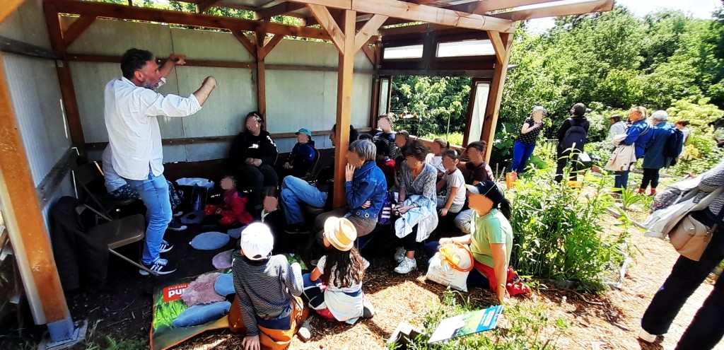 Une quinzaine de personnes assise sous l'abri, un conteur est debout et lève les bras en racontant une histoire. Derrière, un petit groupe debout dans le jardin.